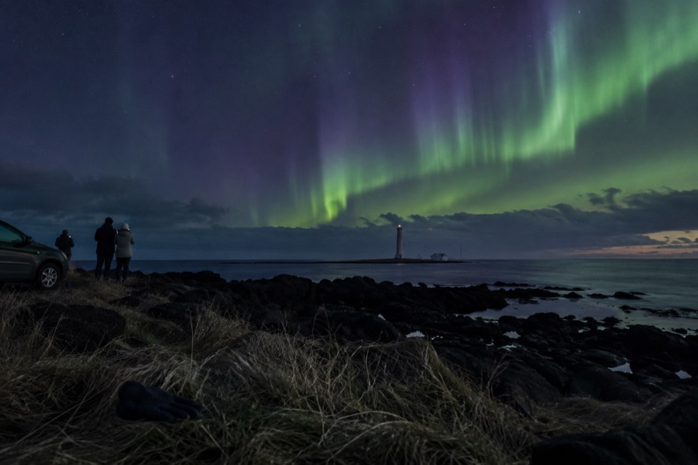Green and purple northern lights dance above a dark shoreline with a lighthouse in the distance.