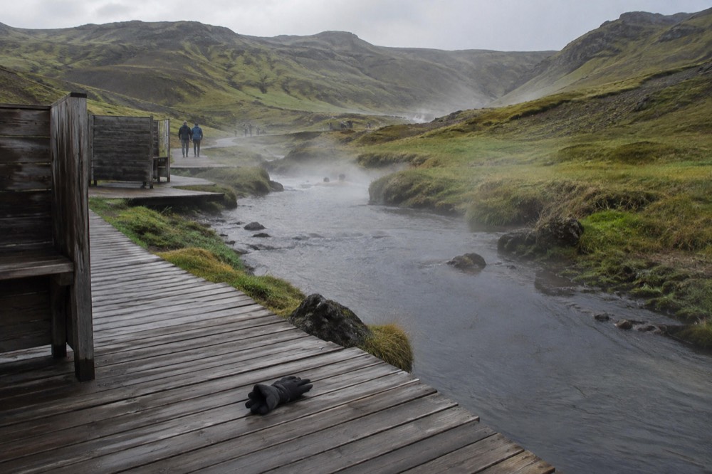 A steaming hot river winding through a green valley with boardwalks and simple wooden screens, no people bathing, the landscape misty and untouched.