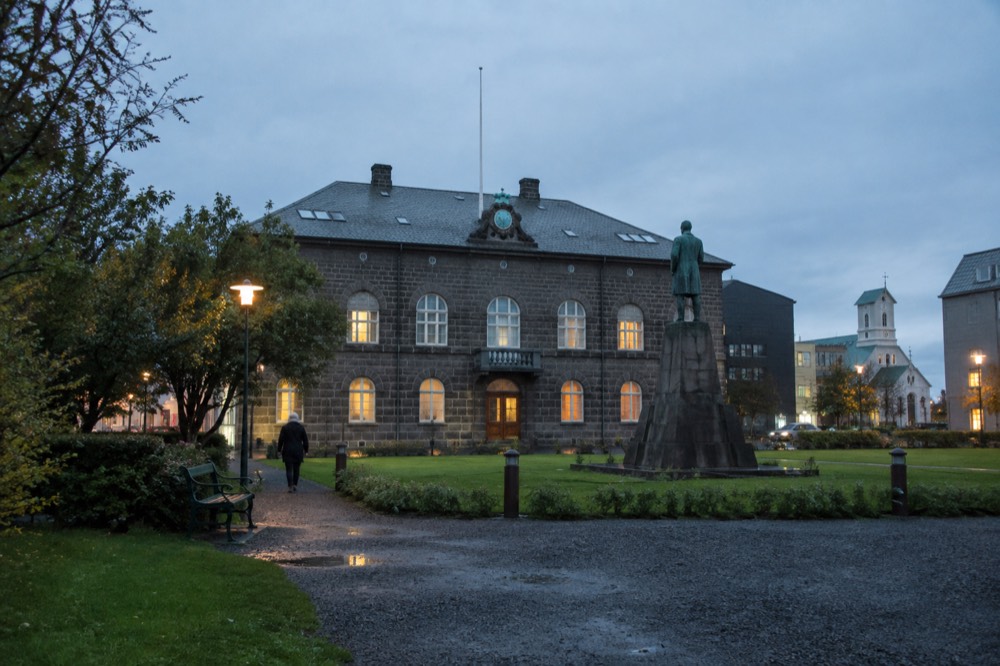 A stone parliament building with a small statue in front, beside a public square with grass and benches