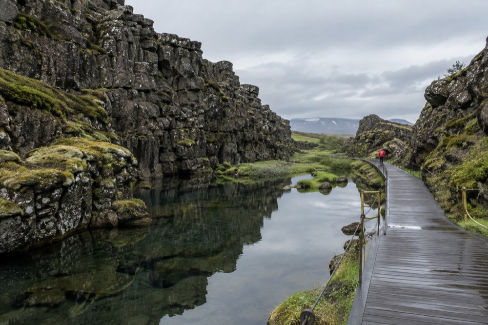 A rift valley path between tall rock walls with a wooden walkway beside still water