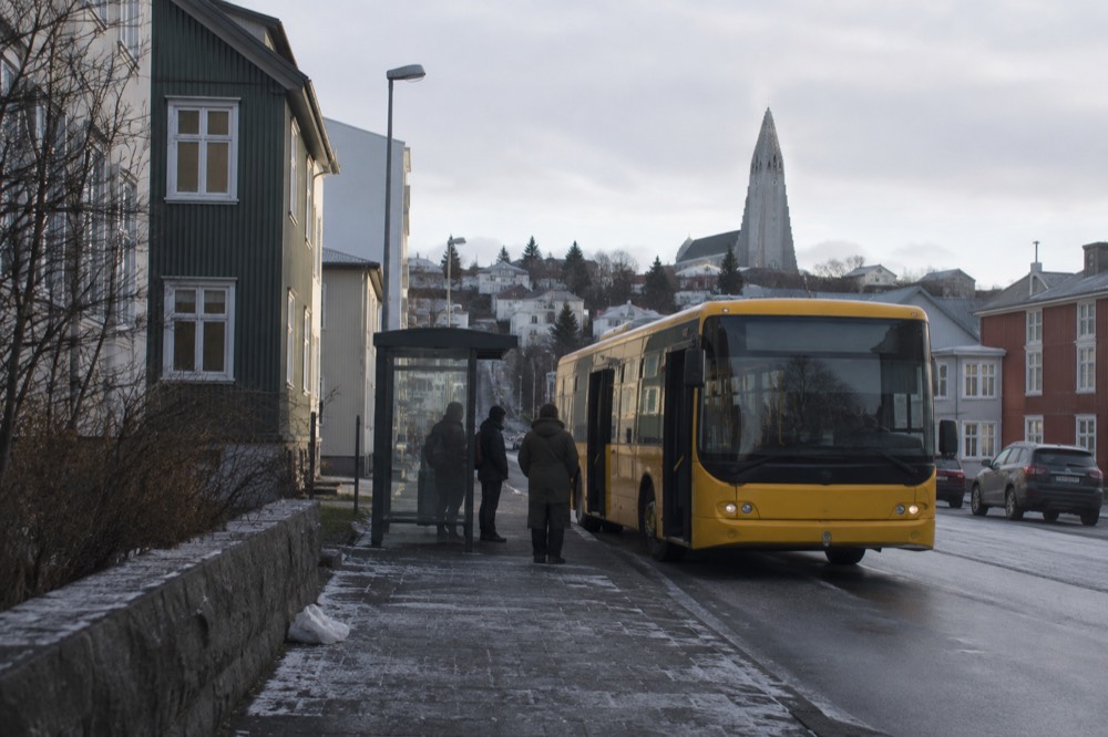 A city bus at a stop on a Reykjavik street with low buildings and a hill behind