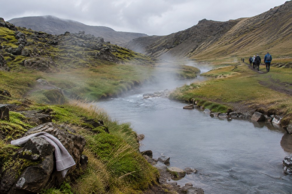 Steam rising from a river running through a green valley with hikers on a path