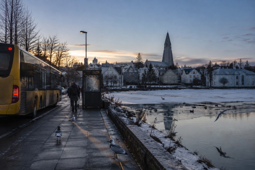 A city bus at a stop beside a pond with a church spire in the distance