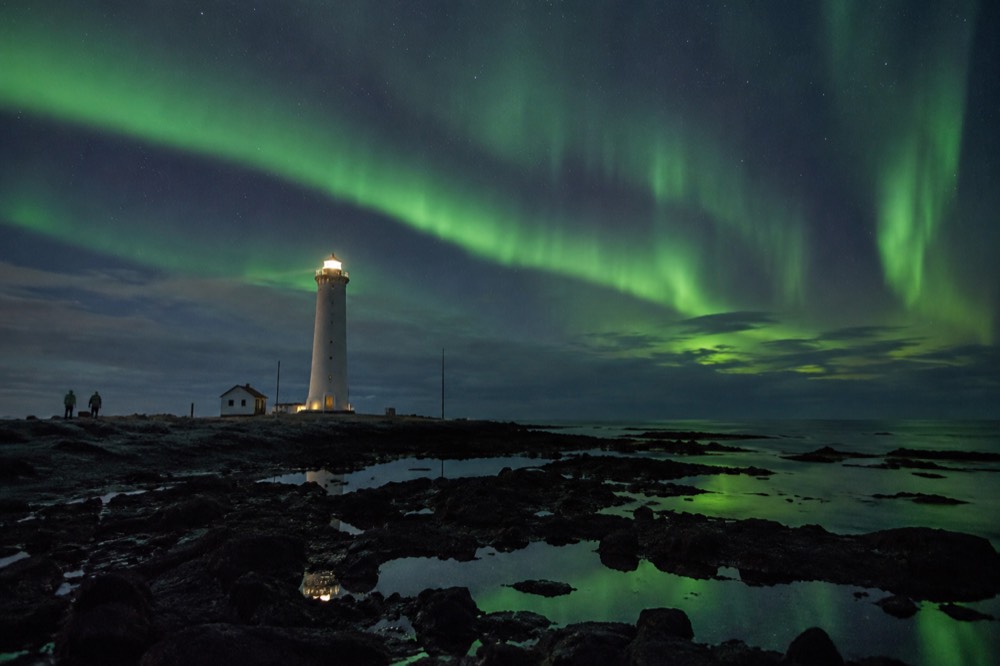 Green aurora bands over a dark shoreline with a lighthouse silhouette