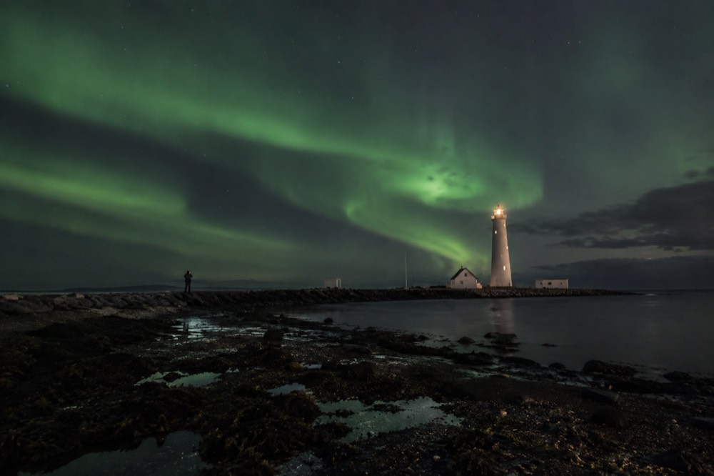 Green aurora arcs over a dark lighthouse and still water