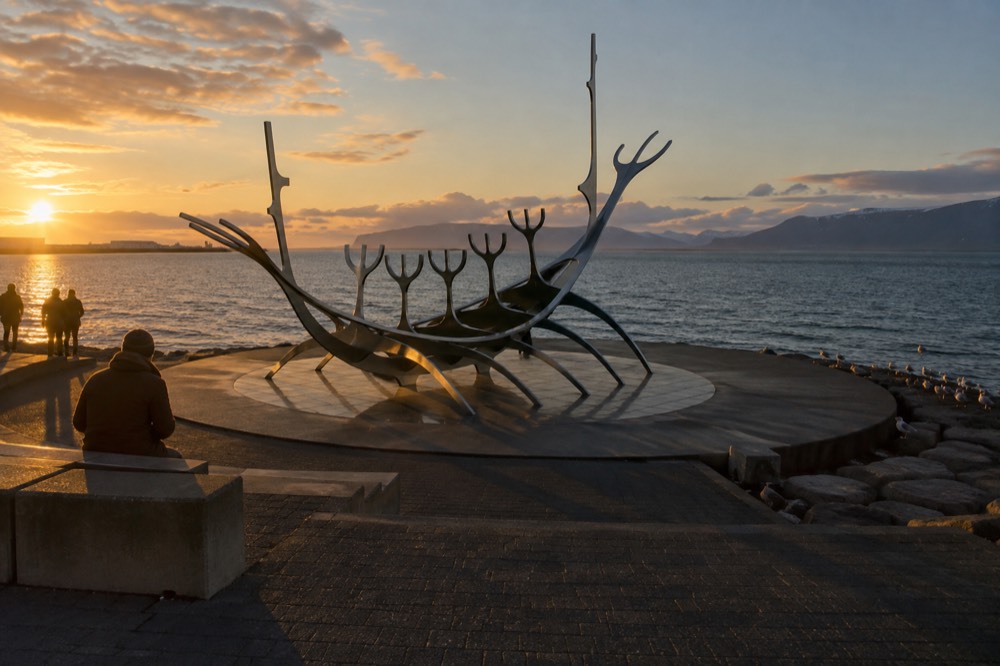 A steel sail-shaped sculpture on a seaside promenade at golden hour