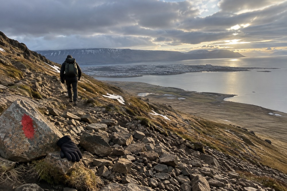 Hiker on a rocky trail with a mountain ridge above a fjord