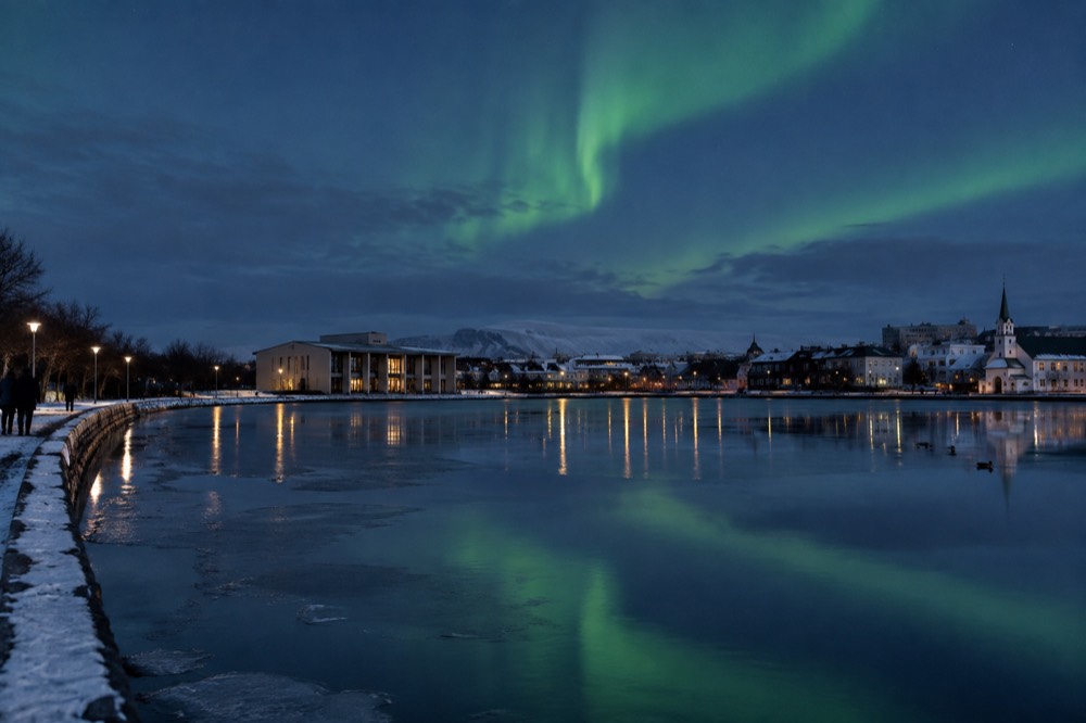 Aurora reflected over a calm pond with city buildings dark on the horizon